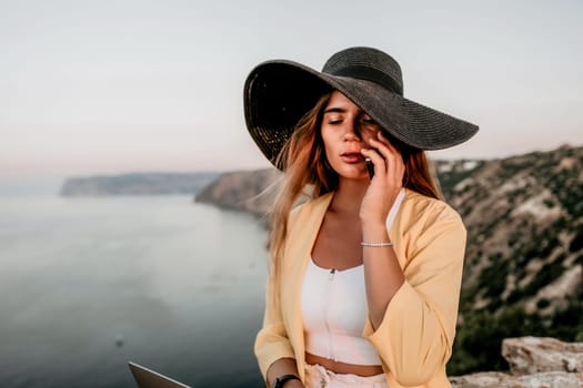 Successful business woman in yellow hat working on laptop by the sea. Pretty lady typing on computer at summer day outdoors. Freelance, travel and holidays concept.