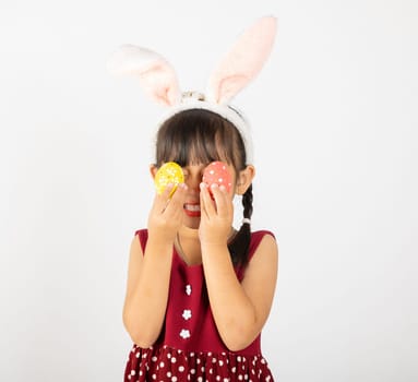 Happy Easter Day. Smile Asian little girl wearing easter bunny ears holding colorfull eggs closes eyes with testicles isolated on white background with copy space, Happy child in holiday