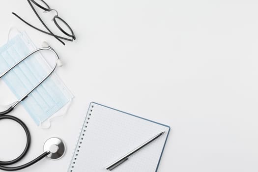 Doctor desktop top view. Spiral notebook with pen and glasses on white background. Stethoscope with syringe and gauze bandage at the workplace of medical worker. Hospital treatment concept, flat lay.