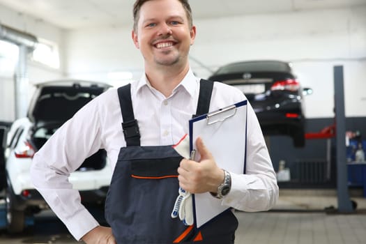 Portrait of caucasian auto mechanic with clipboard of maintenance documents and checking car damage in auto garage. Car repair assistant transport business and after sales service concept