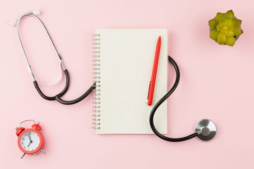 Spiral notepad with a pen on a pink background. Top view. Stethoscope with alarm clock on the doctor table. Flat lay.