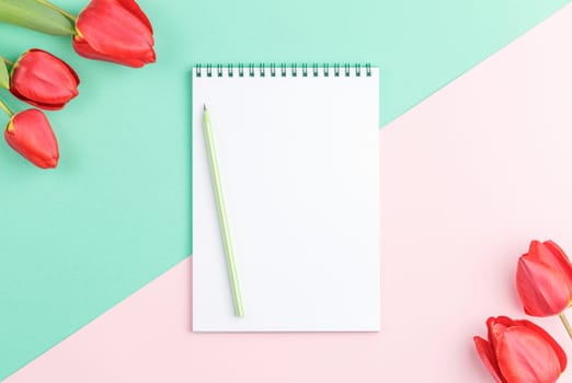 Open spiral notebook with a pencil and red tulip flowers on a green background. Top view. Spring concept.