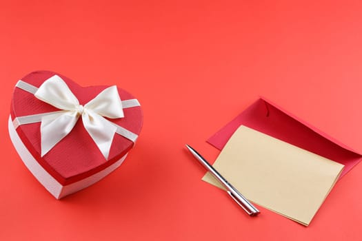 Gift box in the form of a heart with a fabric bow on a red background. Festive concept. Envelope with a pen on the table.