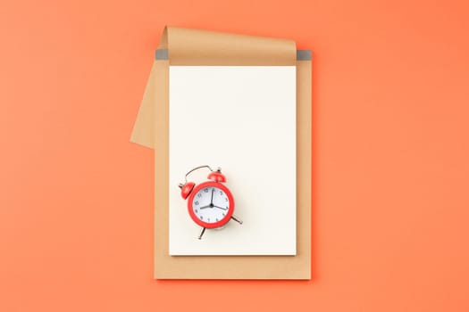 Craft album with white sheets and red alarm clock on orange isolated background top view.