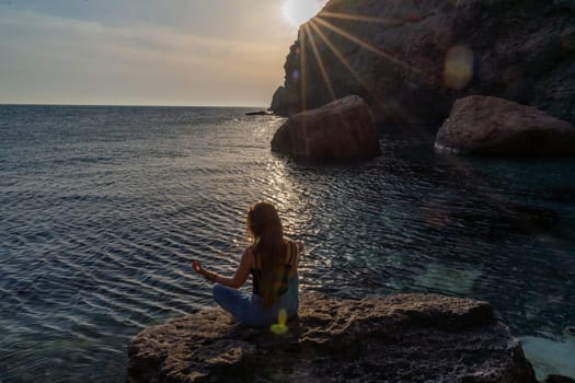 Woman tourist enjoying the sunset over the sea mountain landscape. Sits outdoors on a rock above the sea. She is wearing jeans and a blue hoodie. Healthy lifestyle, harmony and meditation.