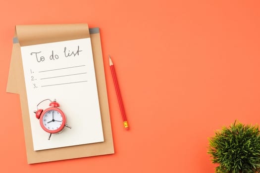 Craft notebook with white sheets, red alarm clock, green leaves plant and pencil on orange isolated background top view.