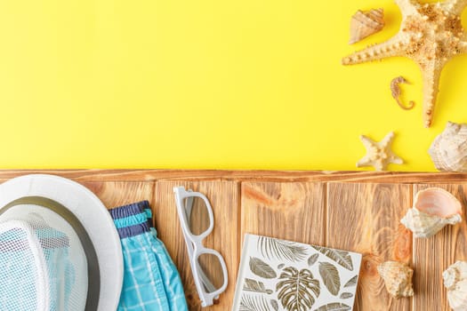 Beach hat with shorts and sunglasses on wooden background with yellow copy space. Flat lay. Summer vacation concept. Starfish and seashells. Top view.