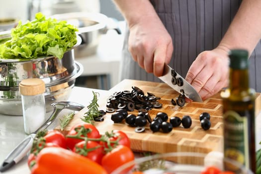 Male cook cuts black olives in kitchen. Process of preparing vegetarian salad concept