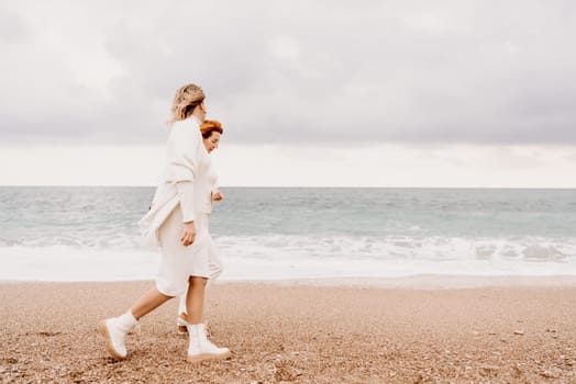 Women sea walk friendship spring. Two girlfriends, redhead and blonde, middle-aged walk along the sandy beach of the sea, dressed in white clothes. Against the backdrop of a cloudy sky and the winter sea. Weekend concept