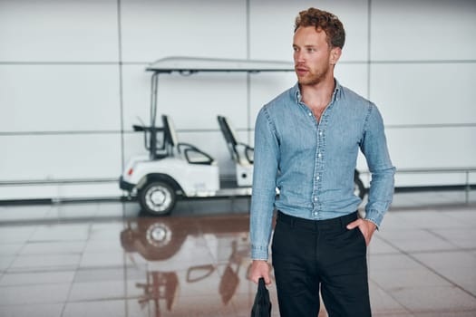 Against vehicle. Young man in formal wear is in modern airport. Conception of vacation.