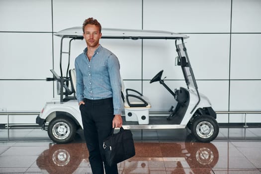 Against vehicle. Young man in formal wear is in modern airport. Conception of vacation.