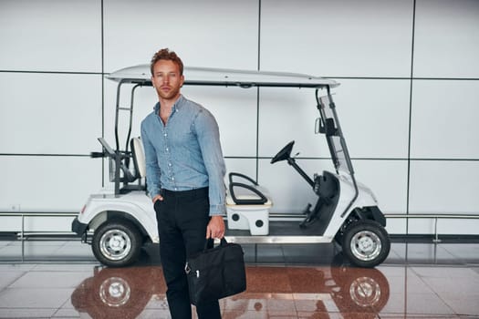 Against vehicle. Young man in formal wear is in modern airport. Conception of vacation.