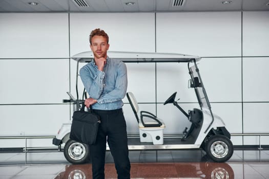 Against vehicle. Young man in formal wear is in modern airport. Conception of vacation.