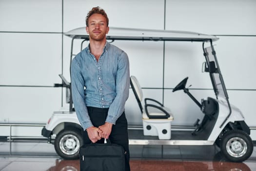 Against vehicle. Young man in formal wear is in modern airport. Conception of vacation.