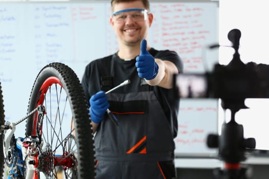 Male blogger holds thumbs up and repairs bicycle remotely. Bike maintenance and live streaming