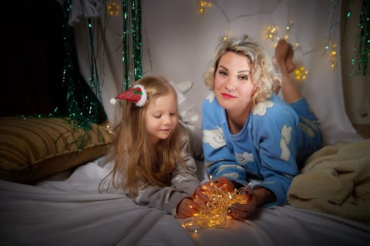 Cute mother and daughter in pajamas lie on soft blanket and having fun in room with Christmas garlands and white background. Tradition of decorating house for holiday. Happy childhood and motherhood