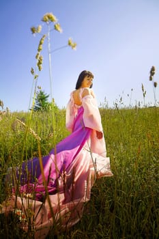 Beautiful girl in lush pink ball gown in green field during blooming of flowers and blue sky on background. Model posing on nature landscape as princess from fary tale