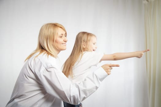 Portrait of a blonde mother and daughter who having communicate and play on white background. Mom and little girl models pointing his finger forward. The concept of family unity