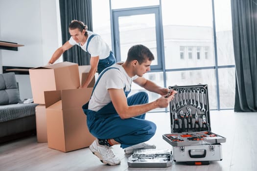 Uses tools from case. Two young movers in blue uniform working indoors in the room.