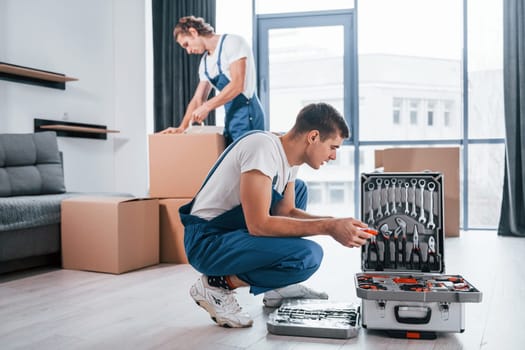 Uses tools from case. Two young movers in blue uniform working indoors in the room.