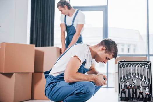 Uses tools from case. Two young movers in blue uniform working indoors in the room.