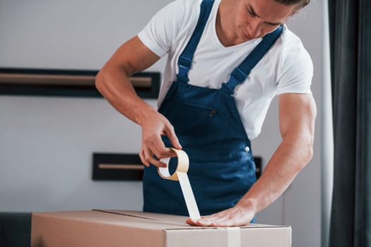 Packaging the box. Young male mover in blue uniform works indoors in the room.