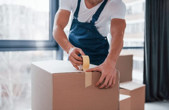 Packaging the box. Young male mover in blue uniform works indoors in the room.