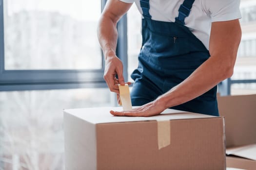 Packaging the box. Young male mover in blue uniform works indoors in the room.