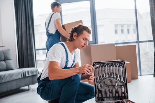 Uses tools from case. Two young movers in blue uniform working indoors in the room.