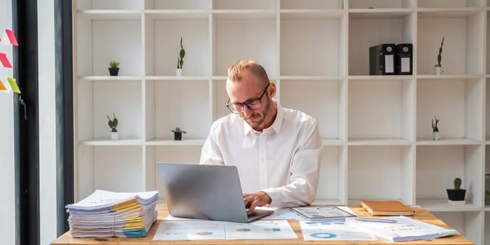 Young business man wearing glasses using laptop, typing on keyboard, writing email or message, sitting in office.