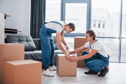 Packaging the box. Two young movers in blue uniform working indoors in the room.