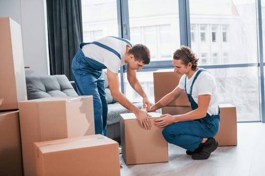 Packaging the box. Two young movers in blue uniform working indoors in the room.