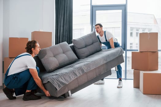 Carrying heavy sofa. Two young movers in blue uniform working indoors in the room.