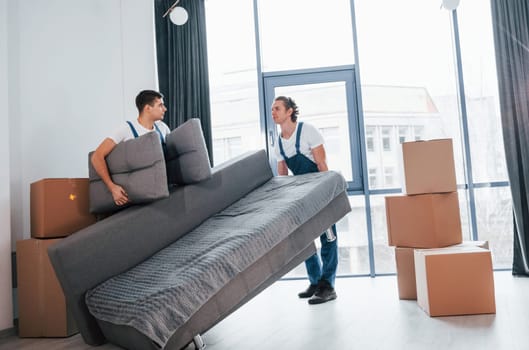 Carrying heavy sofa. Two young movers in blue uniform working indoors in the room.