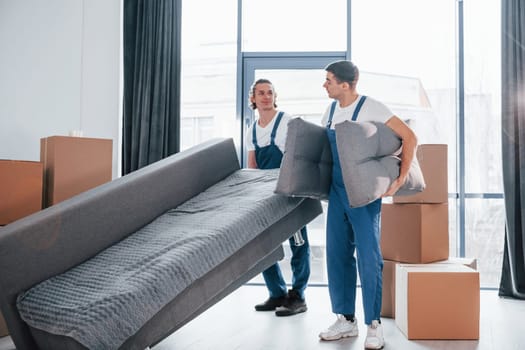 Carrying heavy sofa. Two young movers in blue uniform working indoors in the room.
