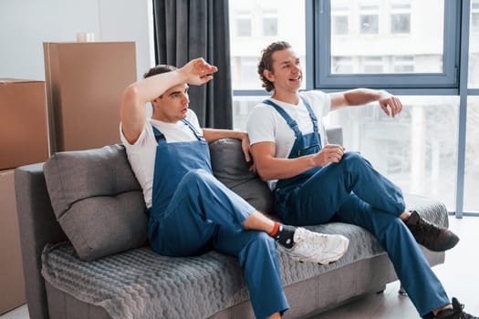 Sitting on the sofa. Tired and taking break. Two young movers in blue uniform working indoors in the room.