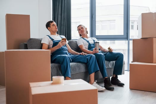 Sitting on the sofa. Tired and taking break. Two young movers in blue uniform working indoors in the room.