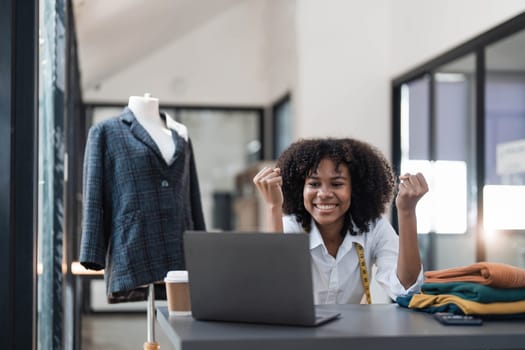 Millennial Asian young professional female designer dressmaker businesswoman with measuring tape on neck sitting smiling hold fists up celebrating together in tailor workshop..