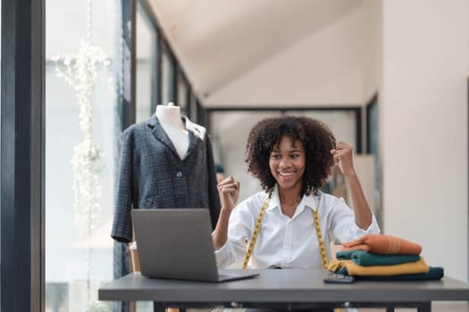 Millennial Asian young professional female designer dressmaker businesswoman with measuring tape on neck sitting smiling hold fists up celebrating together in tailor workshop..