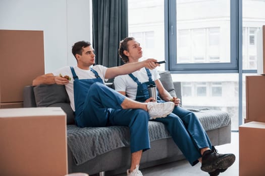 Sitting on the sofa. Tired and taking break. Two young movers in blue uniform working indoors in the room.