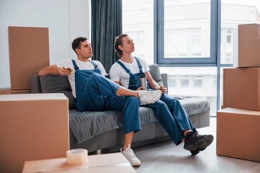 Sitting on the sofa. Tired and taking break. Two young movers in blue uniform working indoors in the room.