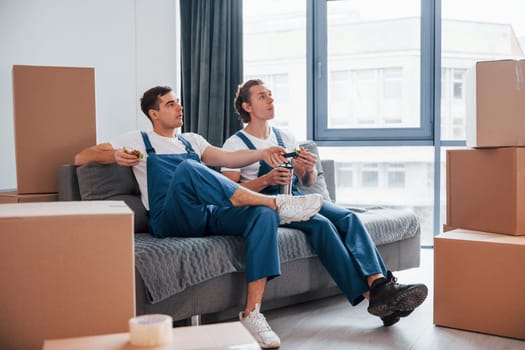 Sitting on the sofa. Tired and taking break. Two young movers in blue uniform working indoors in the room.