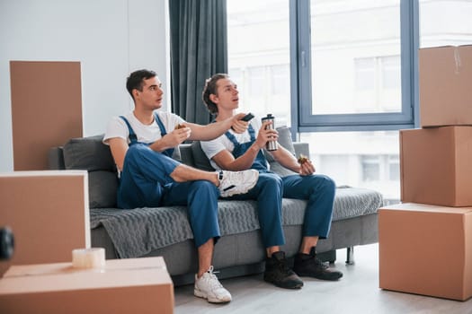 Sitting on the sofa. Tired and taking break. Two young movers in blue uniform working indoors in the room.