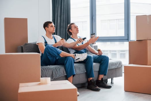 Sitting on the sofa. Tired and taking break. Two young movers in blue uniform working indoors in the room.