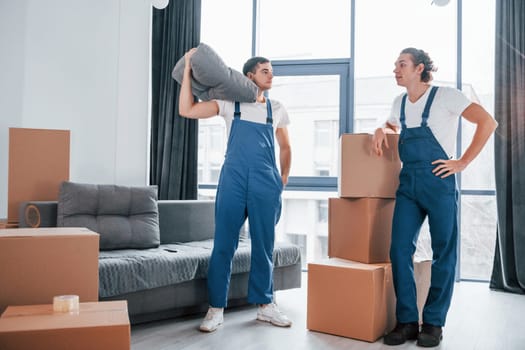 Busy day. Two young movers in blue uniform working indoors in the room.