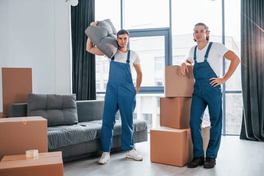 Busy day. Two young movers in blue uniform working indoors in the room.