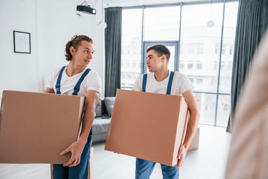 Busy day. Two young movers in blue uniform working indoors in the room.