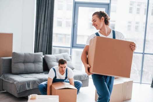 Heavy boxes. Two young movers in blue uniform working indoors in the room.