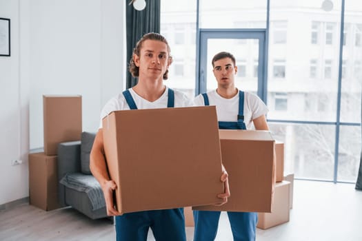 With boxes in hands. Two young movers in blue uniform working indoors in the room.