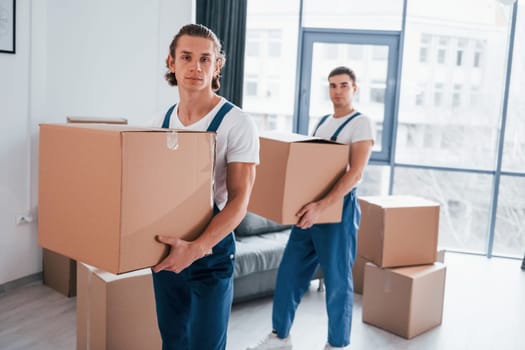 With boxes in hands. Two young movers in blue uniform working indoors in the room.